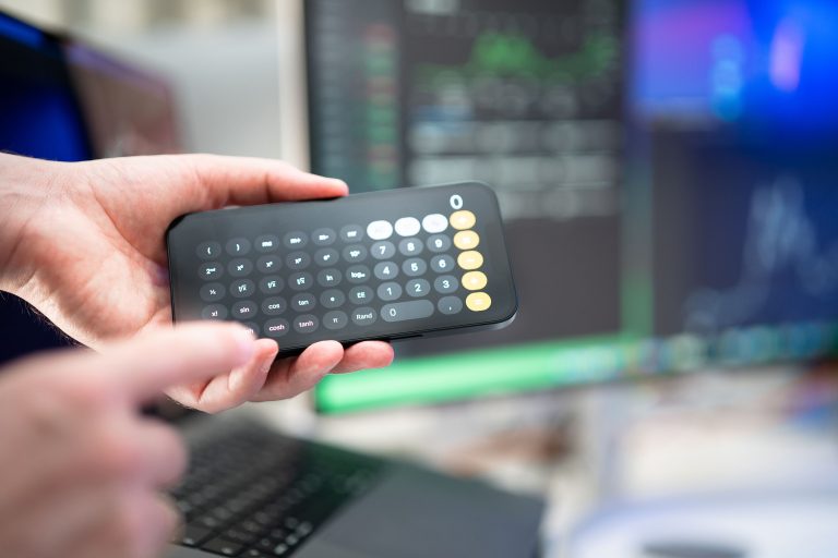 How Founders Should Calculate Product Pricing from Real Costs and Data 2 Close-up of hands using a smartphone calculator amidst computer screens in an office