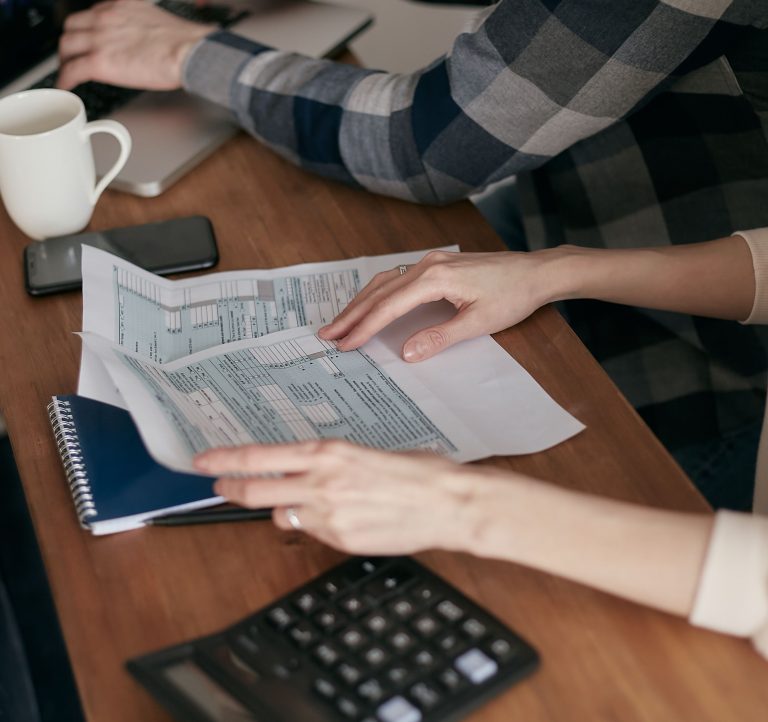 Close-up of hands organizing tax forms on a desk with a calculator, laptop, and notebook