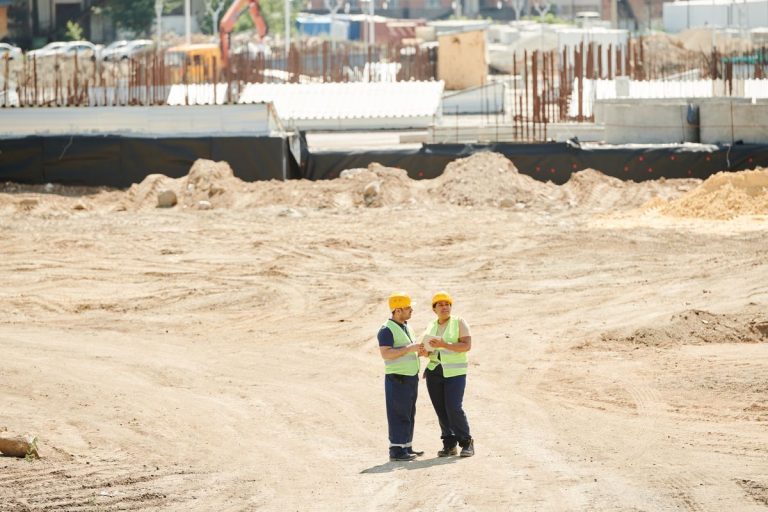 Two construction workers in safety vests talk on a building site
