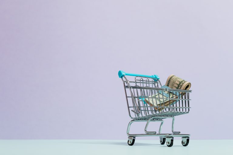 A close-up of a mini shopping cart with rolled cash on a pastel background