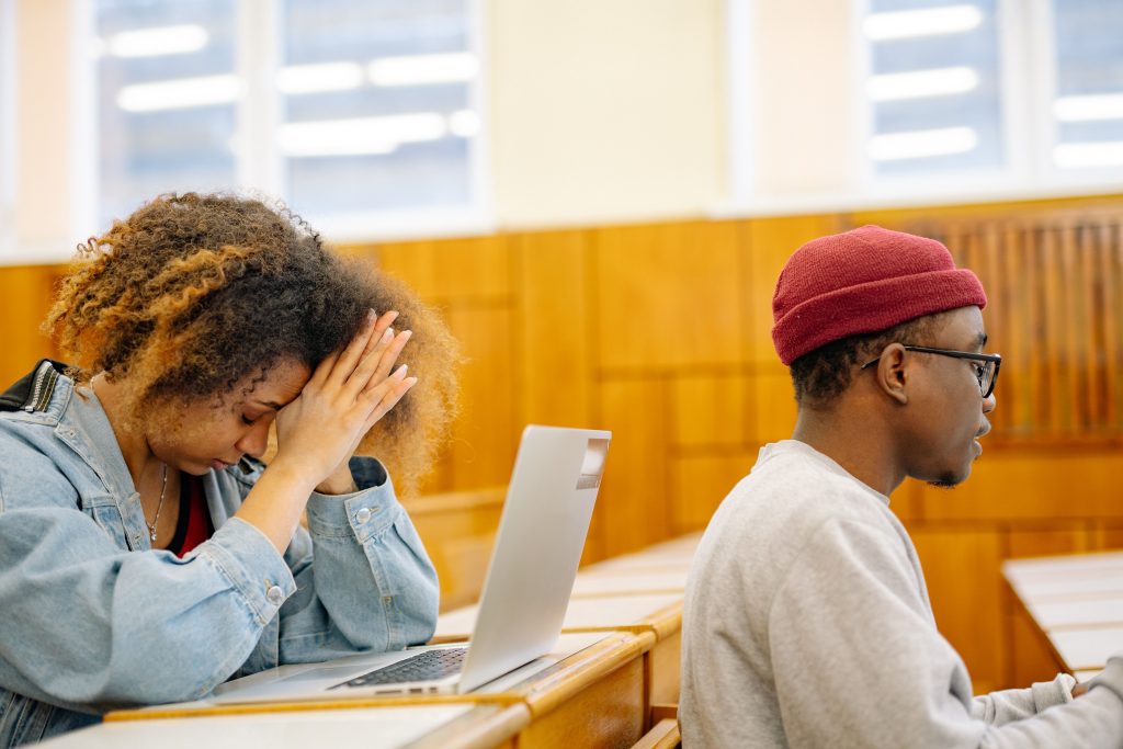 Two students working on laptops in a classroom, showing academic stress