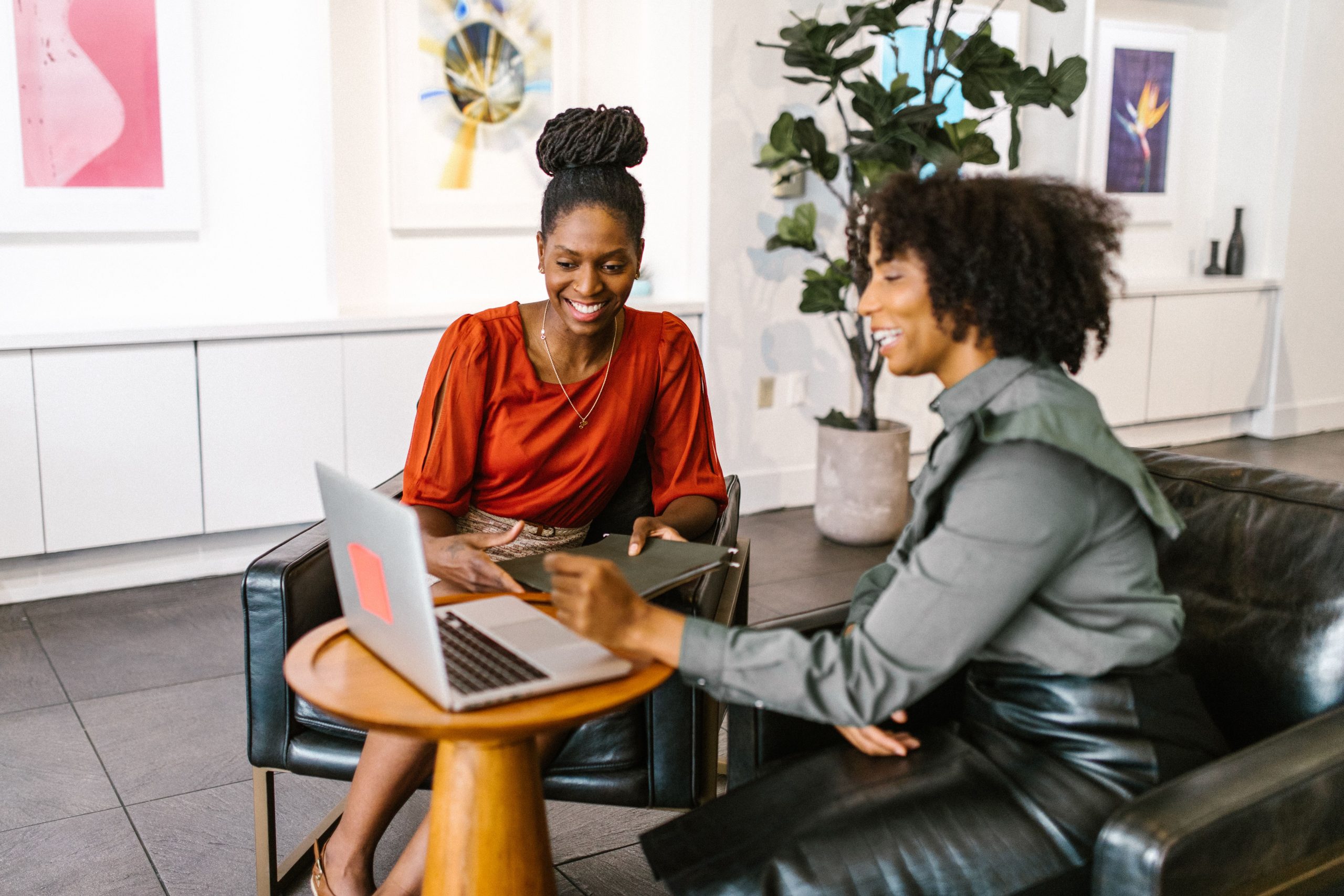 Two women discuss work beside a laptop in an office lounge